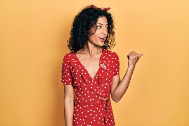 Yazlık elbise giyen, gülümseyen, gülümseyen ve başparmağıyla yan tarafı gösteren genç Latin kız.. 