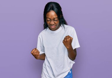 Genç Afro-Amerikalı kadın, gündelik beyaz tişört giyiyor çok mutlu ve heyecanlı, kazanan jest yapıyor, kolları havada, gülümsüyor ve başarı için bağırıyor. kutlama konsepti. 