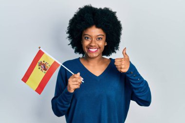Genç Afrikalı Amerikalı kadın elinde İspanya bayrağı tutarken mutlu ve pozitif gülümsüyor, başparmağıyla mükemmel ve onay işareti yapıyor. 