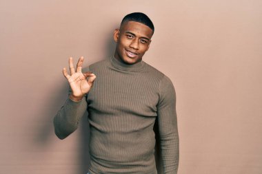 Balıkçı yaka kazak giyen genç siyahi adam gülümsüyor ve el ve parmaklarıyla 