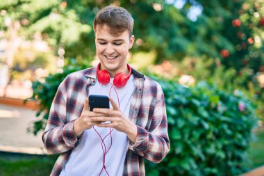 Parkta akıllı telefon ve kulaklık kullanarak gülümseyen beyaz bir genç..