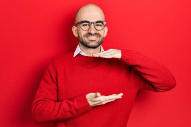 Young bald man wearing casual clothes and glasses gesturing with hands showing big and large size sign, measure symbol. smiling looking at the camera. measuring concept. 