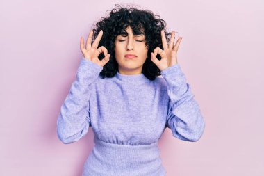 Orta Doğulu genç bir kadın günlük kıyafetler giyip rahatlıyor ve gözleri kapalı gülümsüyor parmaklarıyla meditasyon hareketi yapıyor. yoga konsepti. 