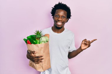 Genç Afro-Amerikalı adam elinde ekmek ve bakkaliyeyle kağıt torba tutarken mutlu bir şekilde elini ve parmağını kenarı işaret ediyor. 