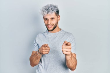 Modern boyalı saçlı, sıradan gri tişörtlü, neşeli ve komik yüzlü genç İspanyol adam kamerayı işaret ediyor. iyi enerji ve titreşimler. 