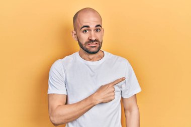 Young bald man wearing casual white t shirt pointing aside worried and nervous with forefinger, concerned and surprised expression 