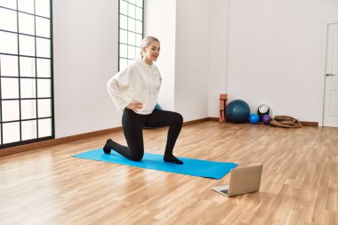 Çevrimiçi yoga dersinde spor merkezinde dizüstü bilgisayar kullanarak mutlu gülümseyen genç sarışın kız..