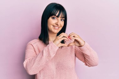 Gündelik giysiler giyen genç İspanyol kadın. Sevgiyle gülümsüyor. Kalp sembolü ve şekli elleriyle gösteriyor. romantik kavram. 