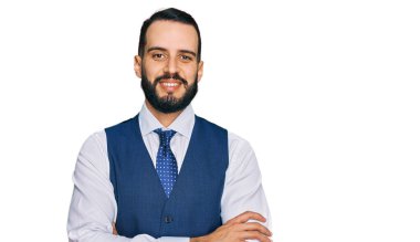 Sakallı, iş yeleği giyen, gülümseyen, kollarını kavuşturup kameraya bakan genç bir adam. pozitif kişi. 