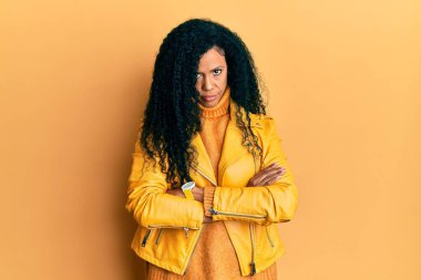 Orta yaşlı Afro-Amerikalı kadın yün kazak ve deri ceket giyiyor şüpheci ve gergin, yüzünde çapraz kollu tasvip etmeyen bir ifade var. negatif kişi. 
