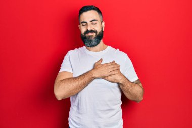 Gündelik beyaz tişört giyen, elleri göğsünde gülümseyen, gözleri kapalı, yüzünde minnet ifadesi olan genç İspanyol bir adam. sağlık kavramı. 