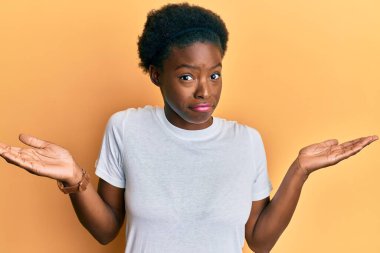 Gündelik beyaz tişört giyen, kafası karışık, kolları açık, hiçbir fikri olmayan, şüpheli yüzlü Afro-Amerikalı genç bir kız.. 