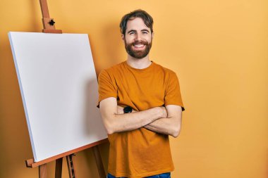 Boyacı sehpasının yanında duran beyaz sakallı adam kollarını kavuşturup kameraya bakarak gülümsüyor. pozitif kişi. 