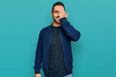 Latin asıllı genç adam gündelik giysiler giyiyor. Bir gözünü eliyle kapatıyor. Yüzünde kendine güvenen bir gülümseme ve sürpriz bir duygu var.. 