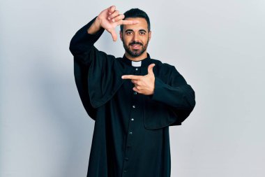 Handsome hispanic man with beard wearing catholic priest robe smiling making frame with hands and fingers with happy face. creativity and photography concept. 