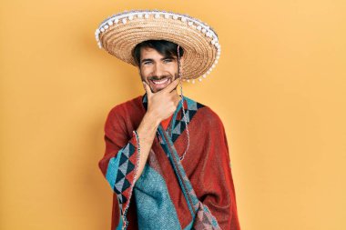Young hispanic man holding mexican hat looking confident at the camera smiling with crossed arms and hand raised on chin. thinking positive. 