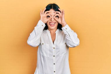 Günlük kıyafetler giyen genç İspanyol kadın dürbün gibi jest yapıyor, gözleri parmaklarından bakıyor. çılgın ifade. 