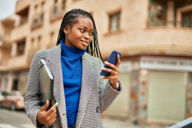 Genç Afrikalı Amerikalı iş kadını şehirde akıllı telefon kullanarak mutlu bir şekilde gülümsüyor..