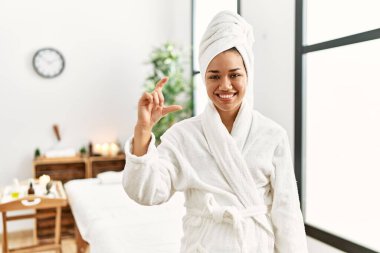 Havlu ve bornoz giymiş genç esmer kadın güzellik merkezinde duruyor gülümsüyor ve kendinden emin el hareketleriyle küçük beden işareti yapıyor parmaklarıyla ve kamerayla bakıyor. ölçüm kavramı. 