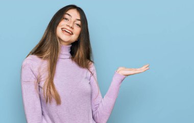 Balıkçı yaka kazak giyen genç ve güzel bir kız neşeli bir şekilde gülümsüyor ve avuç içi kameraya bakıyor.. 