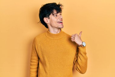 Yakışıklı hippi genç adam, günlük sarı süveter giyiyor, gülümsüyor, gülümsüyor ve başparmağıyla yan tarafa bakıyor.. 