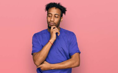 Young african american man with beard wearing casual purple t shirt with hand on chin thinking about question, pensive expression. smiling with thoughtful face. doubt concept. 
