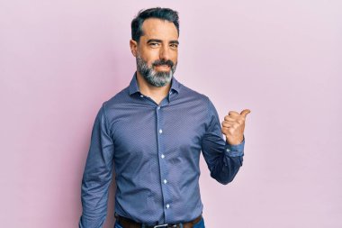 Orta yaşlı, sakallı, gri saçlı, iş kıyafetleri giyen, gülümseyen, gülümseyen ve baş parmağıyla yüzünü gösteren bir adam.. 