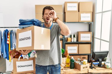 Handsome middle age man holding donations box for charity at volunteer stand doing ok gesture shocked with surprised face, eye looking through fingers. unbelieving expression. 