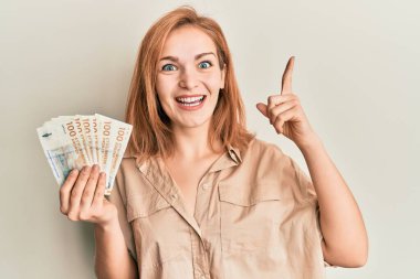 Young caucasian woman holding 100 danish krone banknotes smiling with an idea or question pointing finger with happy face, number one 