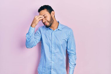 Hispanic man with beard wearing casual blue shirt tired rubbing nose and eyes feeling fatigue and headache. stress and frustration concept. 