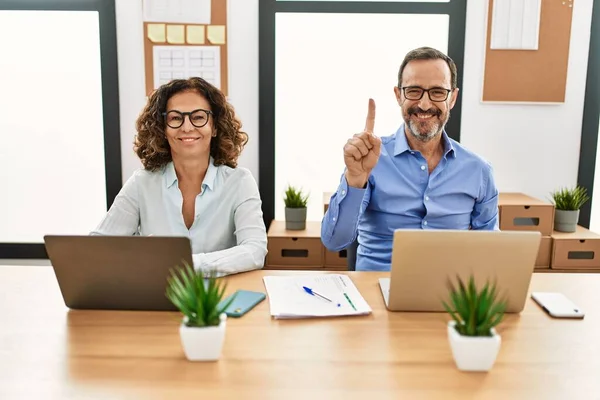 Middle age hispanic woman and man sitting with laptop at the office showing and pointing up with finger number one while smiling confident and happy.