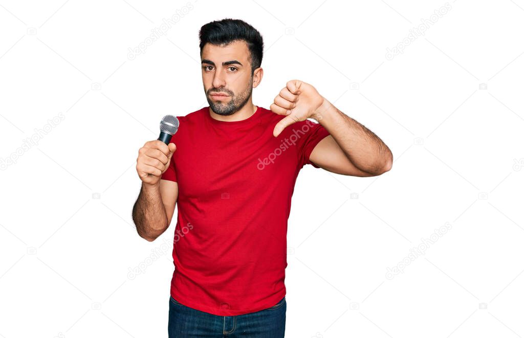 Hispanic man with beard singing song using microphone with angry face, negative sign showing dislike with thumbs down, rejection concept 