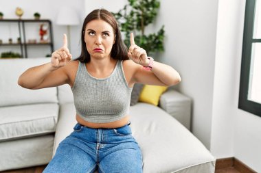 Evdeki kanepede oturan genç esmer kadın üzgün ve üzgün görünüyor, parmaklarıyla yönü gösteriyor, mutsuz ve depresif.. 