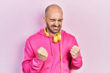 Spor kıyafetleri giymiş, kulaklık takan genç kel bir adam çok mutlu ve heyecanlı bir şekilde kolları havada, gülümseyerek ve başarı için bağırarak kazanan bir jest yapıyor. kutlama konsepti. 