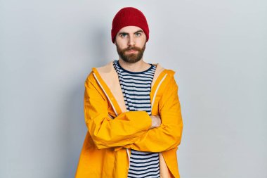 Sakallı, sarı yağmurluk giyen, kuşkucu ve gergin, yüzünde çapraz kollu tasvip etmeyen beyaz bir adam. negatif kişi. 