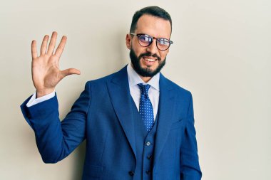 Sakallı, takım elbiseli ve kravatlı genç adam beş numaralı parmaklarını gösterip gösterirken kendinden emin ve mutlu gülümsüyor.. 