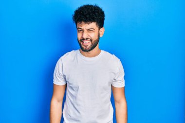 Sakallı, beyaz tişörtlü, seksi bir ifadeyle kameraya bakan, neşeli ve mutlu yüzlü genç bir Arap.. 