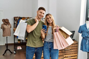 Alışveriş torbaları tutan ve giyim mağazasında akıllı telefon kullanan İspanyol bir çift..