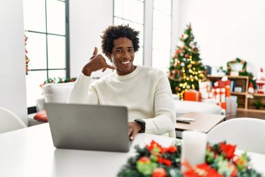 Genç Afro-Amerikalı adam dizüstü bilgisayar kullanıyor Noel ağacının yanında oturuyor gülümsüyor telefonla el ve parmak hareketleri yapıyor telefonda konuşuyor gibi. iletişim kavramları. 