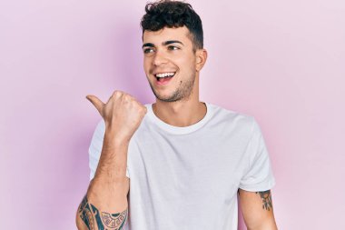 Gündelik beyaz tişört giyen genç İspanyol adam baş parmağını yana kaldırmış, ağzı açık gülümsüyor. 