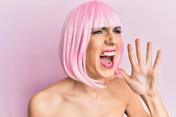 Young man wearing woman make up wearing pink wig shouting and screaming loud to side with hand on mouth. communication concept.