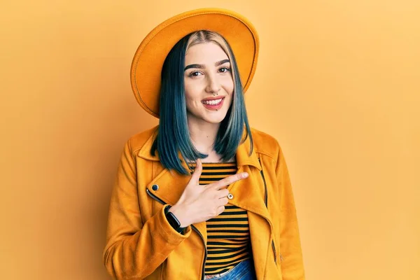 黄色の帽子と革のジャケットを身に着けている若い現代の女の子陽気に笑顔で顔を指します手と指で幸せと自然な表情で側面まで — ストック写真