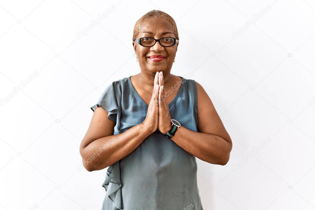 Mature hispanic woman wearing glasses standing over isolated background praying with hands together asking for forgiveness smiling confident. 
