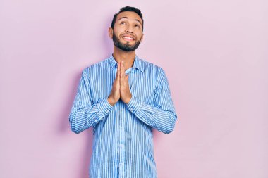 Sakallı, sıradan mavi tişörtlü İspanyol bir adam elleri bağlı yalvarıyor ve dua ediyor. Yüzünde umut ifadesi var. Çok duygusal ve endişeli. Yalvarıyor. 