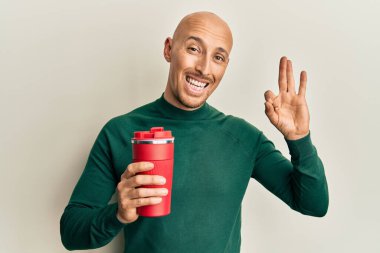 Kel ve sakallı bir adam bir fincan kahve içiyor tamam işareti yapıyor parmaklarıyla, gülümseyerek mükemmel bir sembol oluşturuyor 