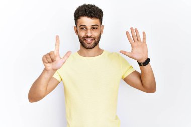Arap genç adam izole edilmiş arka planda duruyor ve yedinciyi parmaklarıyla gösterip işaret ederken kendinden emin ve mutlu gülümsüyor.. 