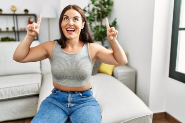 Genç esmer kadın evdeki kanepede oturuyor. Şaşırmış, şaşırmış ve parmaklarıyla işaret ediyor ve kollarını kaldırıyor.. 