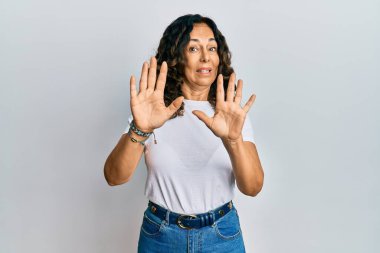 Orta yaşlı İspanyol bir kadın günlük beyaz tişört giyiyor korku ifadesinden korkuyor ve dehşete düşüyor. El kol hareketi yapmayı kes, şok içinde bağırmayı kes. panik konsepti. 