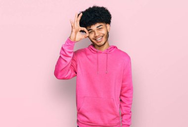 Afro saçlı, sıradan pembe kazak giyen ve gülümseyen Afro Amerikalı genç adam el ve parmaklarıyla tamam işareti yapıyor. başarılı ifade. 