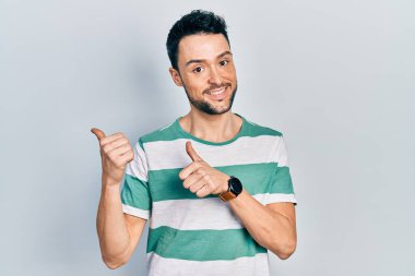 Gündelik kıyafetler giyen genç İspanyol adam arka tarafı işaret ediyor. Elleri havada, kendinden emin bir şekilde gülümsüyor. 